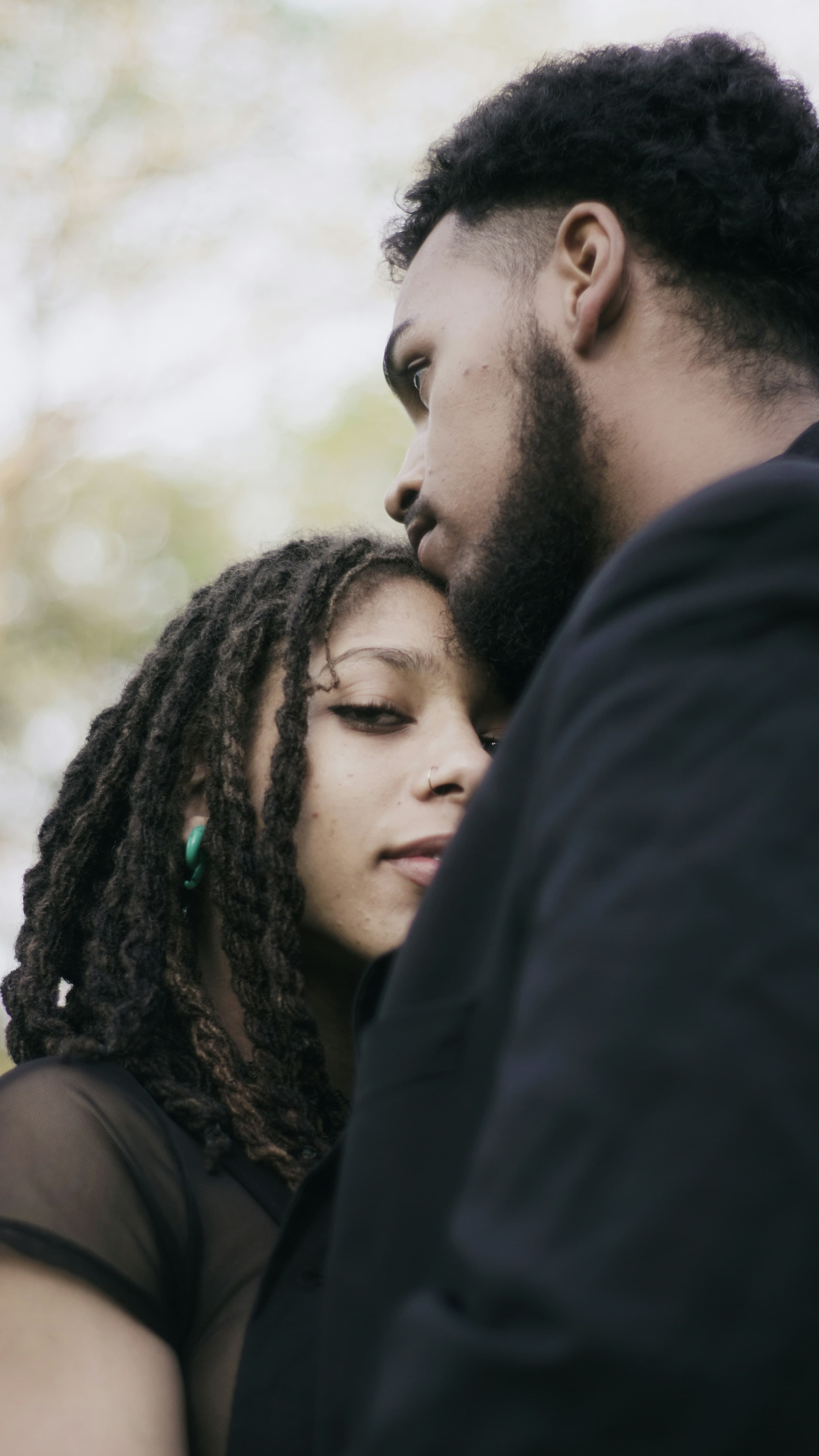 A couple in a quiet embrace outdoors, the man resting his cheek on the woman's forehead